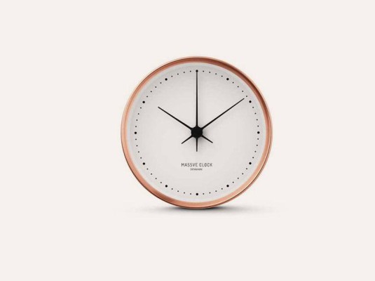The image shows a Massive Clock, a minimalist wall clock with a rose gold colored frame and a white face.  The hands and hour markers are simple and black. The brand name is subtly printed at the center of the clock face.