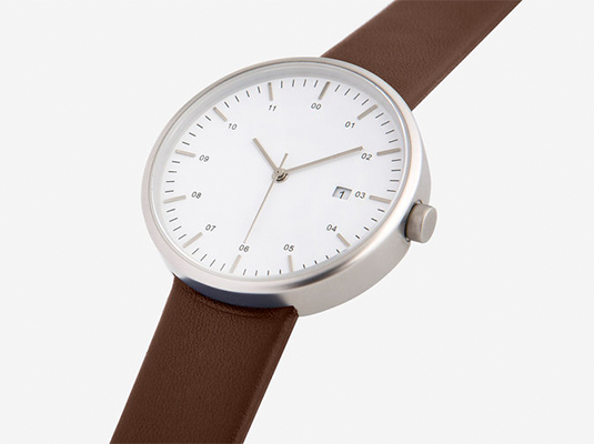 The image shows a close-up view of a wristwatch. The watch has a minimalist design with a round, silver-colored case and a white face. The watch hands and hour markers are simple and understated, and the date window is positioned at three o'clock. The watch strap is made of brown leather.  The overall aesthetic is clean, modern, and sophisticated.