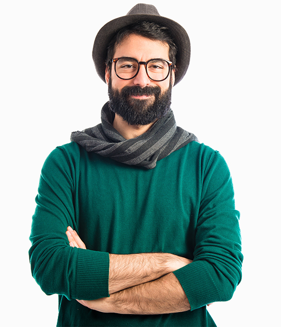 The image shows a man with a beard and glasses, wearing a green sweater, a grey scarf, and a brown hat. His arms are crossed, and he has a calm and self-assured expression on his face. The background is plain white, focusing attention on the subject.  The overall impression is one of quiet confidence and understated style.