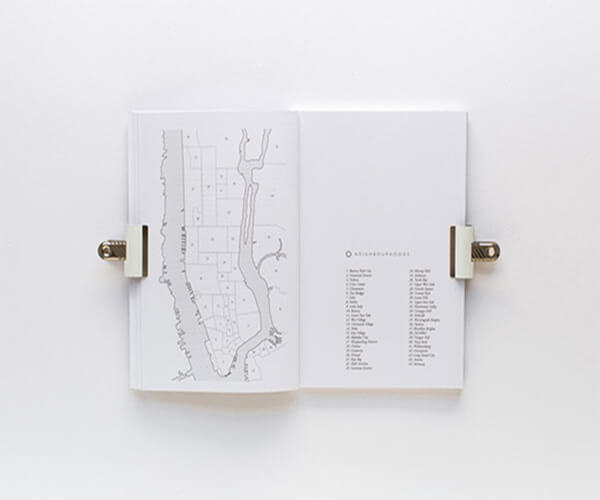 The image shows an open book or booklet lying flat.  The left-hand page features a simplified line drawing map of Manhattan Island, New York City, with numbered neighborhoods. The right-hand page contains a numbered list, presumably corresponding to the numbered areas on the map, listing the names of these neighborhoods.  Two simple, off-white binder clips hold the pages open. The overall aesthetic is clean, minimalist, and monochromatic.