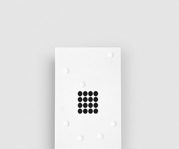 The image shows a minimalist composition.  A white square card or board is the central focus, featuring a small grid of black dots in the center.  Several small, white dots are scattered around the edges of the white card, creating a simple, yet visually interesting contrast. The background is a plain, light gray, further emphasizing the stark simplicity of the main element.