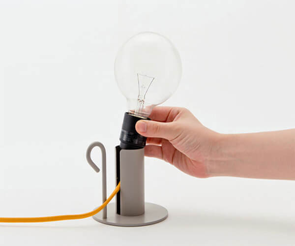 The image shows a hand placing a light bulb into a simple, minimalist table lamp. The lamp has a cylindrical grey base and a small, curved handle. The cord is bright orange.  The overall aesthetic is modern and clean.