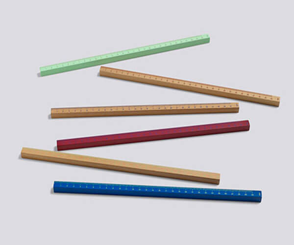 The image shows six rulers of different colors arranged on a light gray surface.  The rulers are thick and appear to be made of wood, with each having a different color of paint or stain: light green, light brown, natural wood, dark red, natural wood, and dark blue.  The rulers are casually arranged, overlapping slightly, and not precisely aligned.
