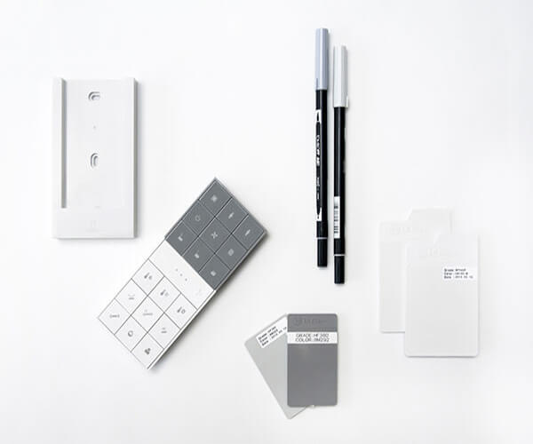 The image shows a minimalist, modern design concept.  It features a white wall-mounted control panel, a grey remote control with multiple buttons, two fine-tip markers (one grey, one black), and several small, rectangular color swatches in varying shades of grey and white.  The overall aesthetic is clean, simple, and suggests a focus on precision and design detailing, possibly related to interior design or home automation.